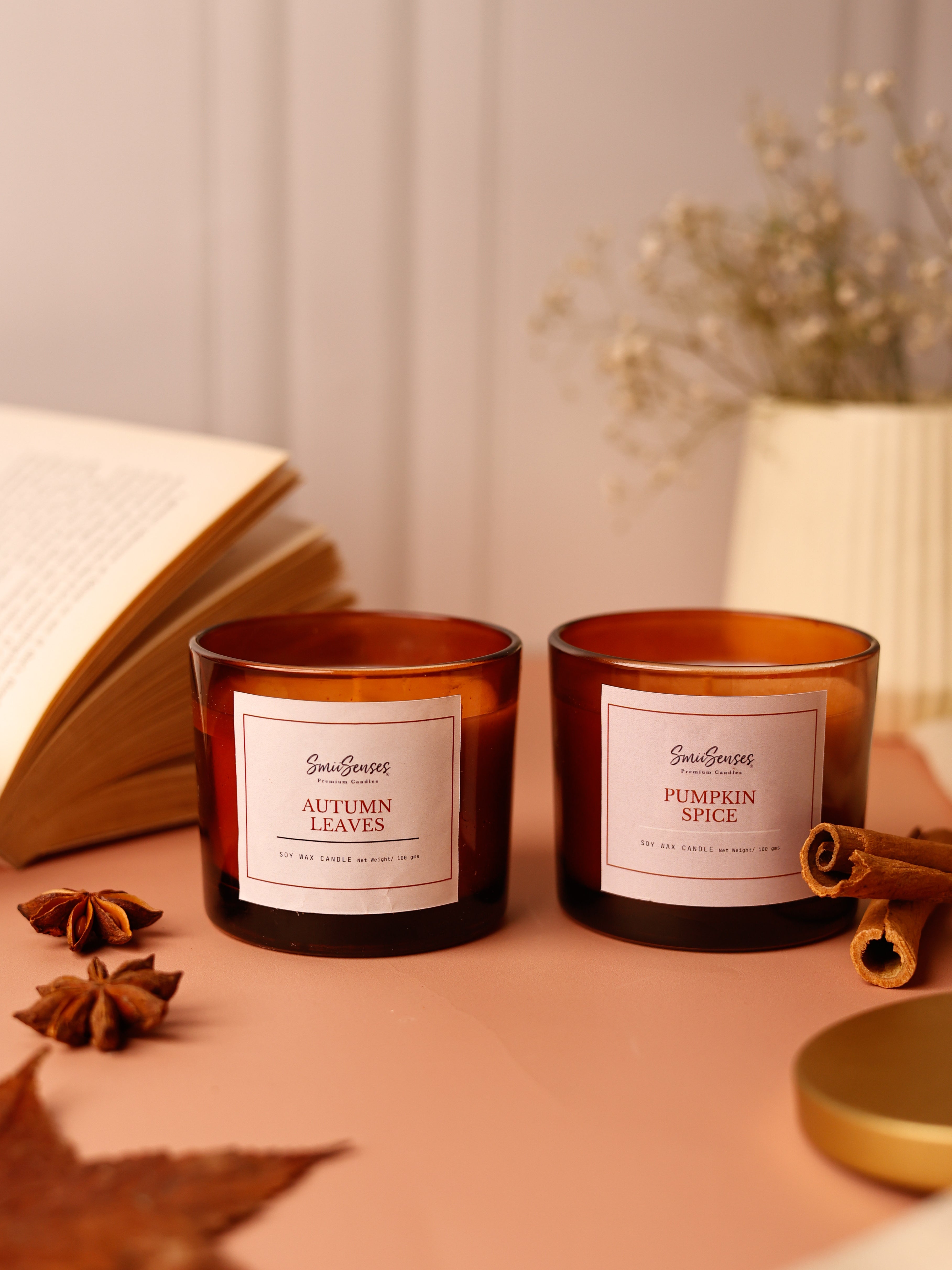 Two melted candles in brown jars with autumn-themed labels, placed on a table with leaves and a book in the background.